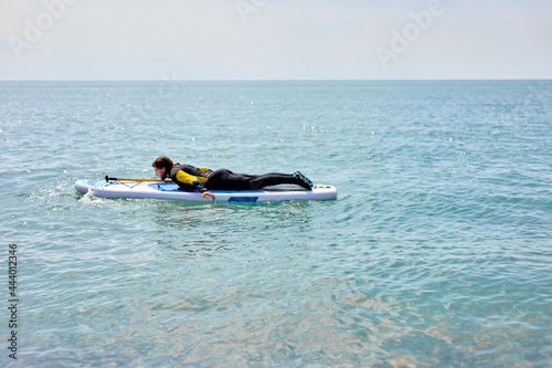 Handsome caucasian male in wetsuit lying on stomach on board while paddleboarding, enjoy paddleboarding alone, at sea on blue water. side view portrait