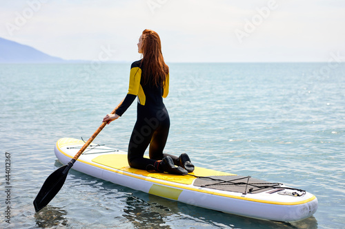 Rear view on fit redhead woman in wetsuit standing paddling away on stand up paddleboarding at sea on blue water. Beautiful slim female is standing on sup surfboard, watersport leisure activity. © Roman