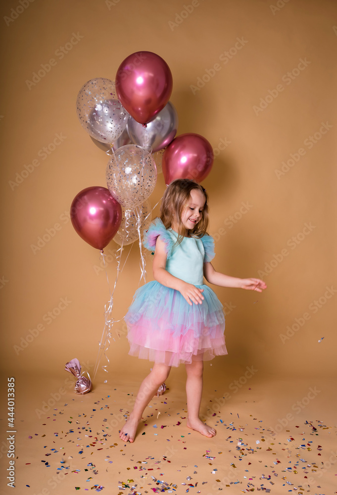 cheerful little girl in a festive dress with a tutu skirt on a beige background with a bunch of inflatable balloons and confetti. Holiday birthday