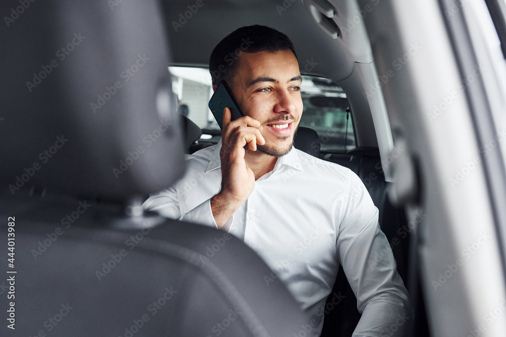 Talks by phone. Young man in white shirt is sitting inside of a modern new automobile Talks by phone. Young man in white shirt is sitting inside of a modern new automobile