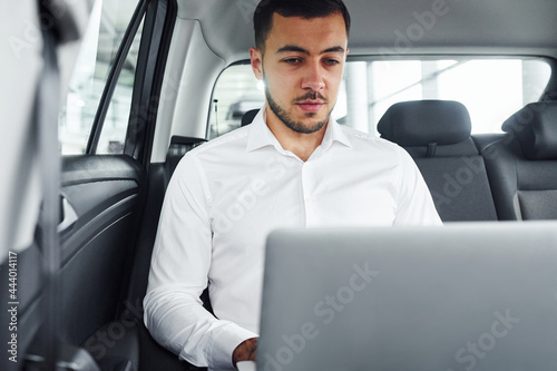 Works by using laptop. Young man in white shirt is sitting inside of a modern new automobile © standret