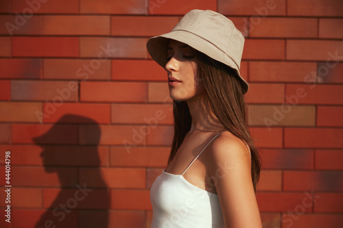 Stylish millennial woman in hat standing near brick wall