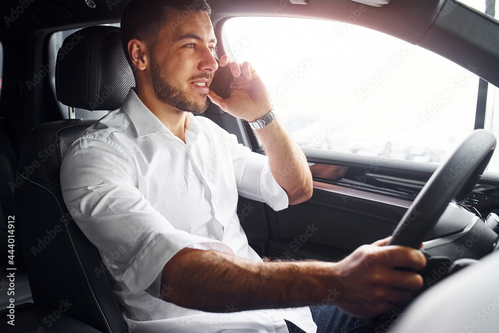 Successful man. Young guy in white shirt is sitting inside of a modern new automobile