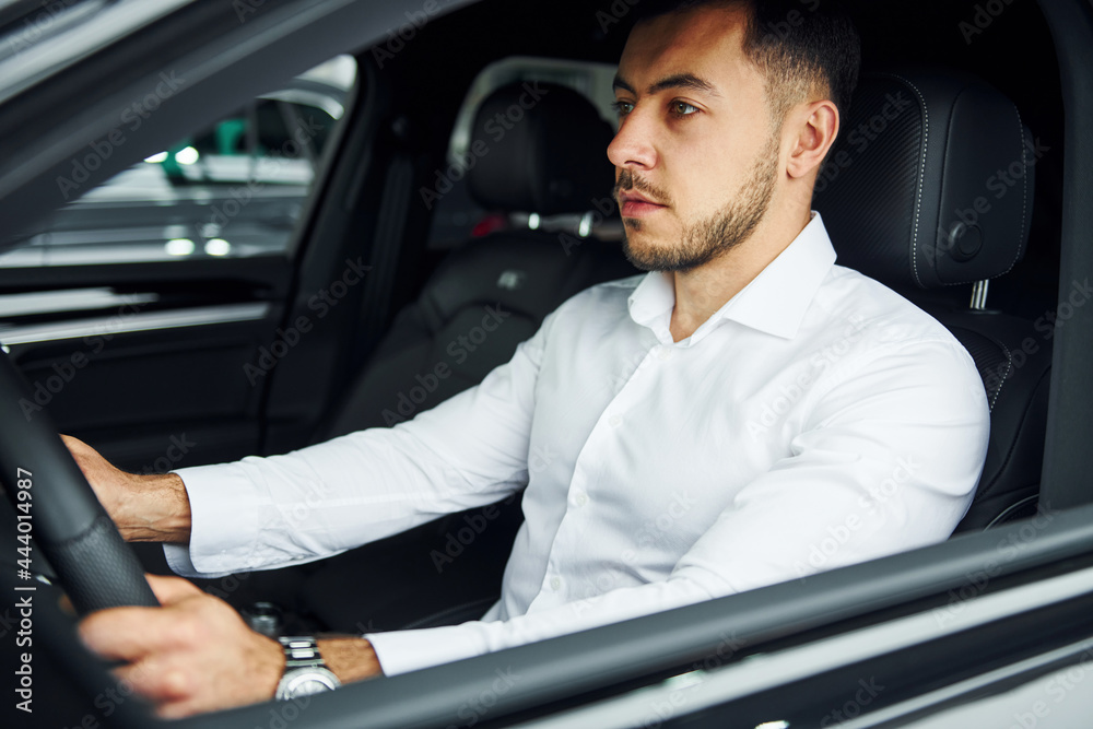 Successful man. Young guy in white shirt is sitting inside of a modern new automobile