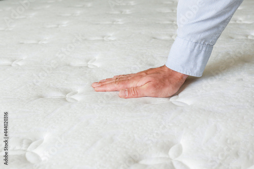 Cropped shot of man testing the firmness of white orthopedic mattress. Male pressing hypoallergenic foam mattresses surface to check its softness. Close up, copy space, top view, background. photo