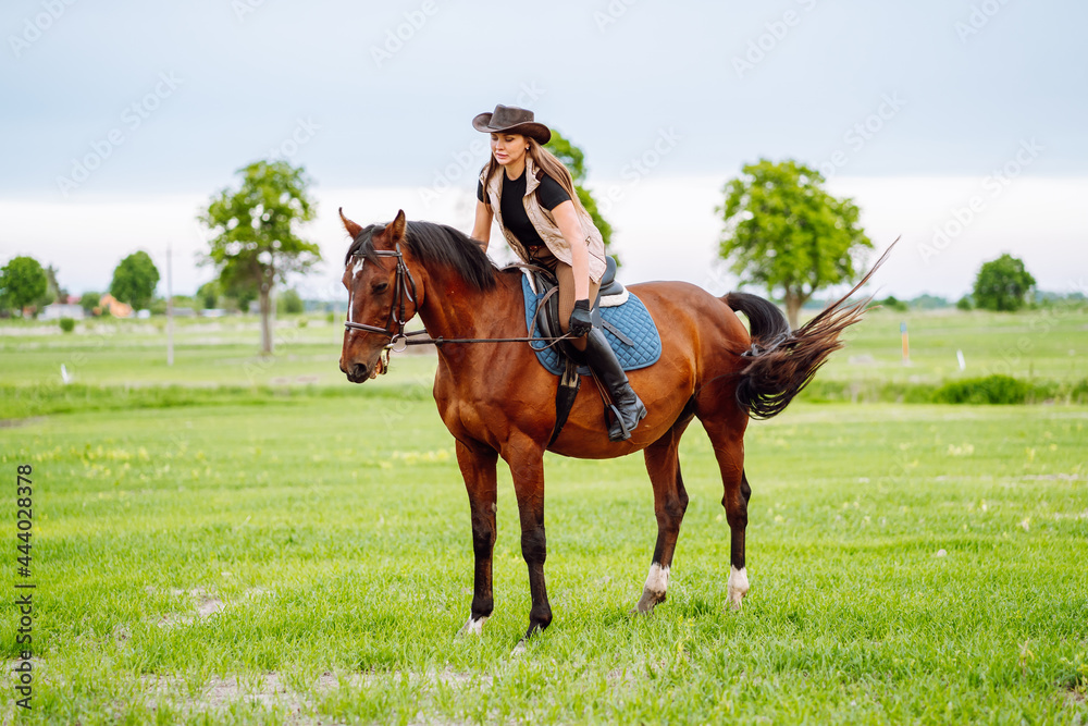 Young woman dressed in riding clothes and hat riding brown horse in green field