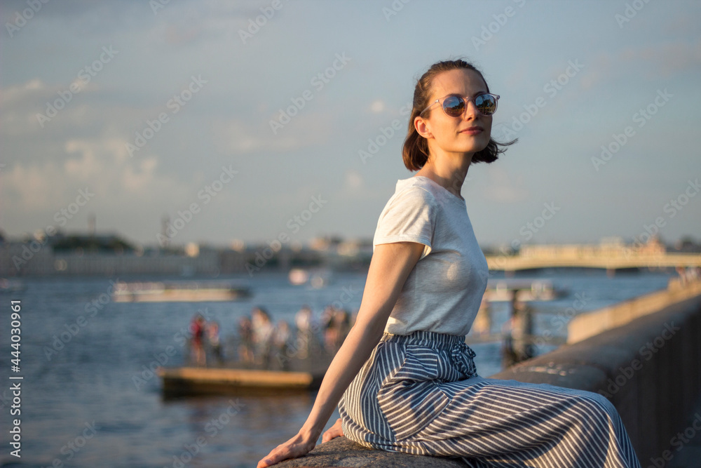 beautiful young woman sitting by the river on the background of the cityscape in sunset