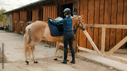 Female jockey saddling up her light brown horse outside the stable. Placing a leather saddle on the back of her horse. The girl wearing a helmet and gloves. Preparing her horse for the competition.