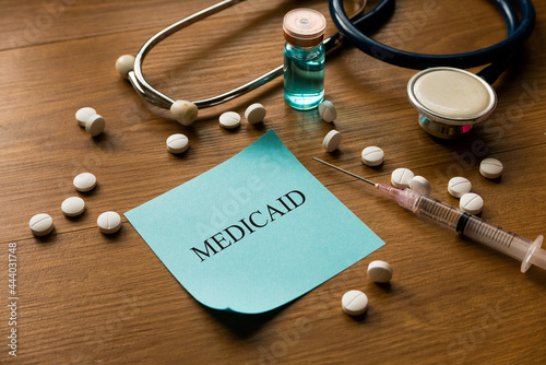 Selective focus of stethoscope, white medical pills, syringe and blue memo note written with Medicaid on wooden background.