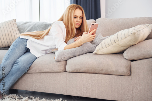 Lying down on sofa. Young woman in white shirt and jeans is at home