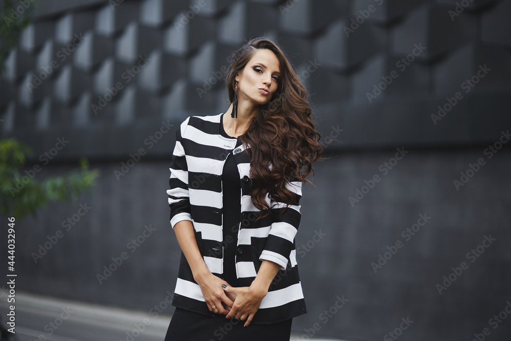 Gorgeous young model woman with perfect dark hair wearing black evening dress and jacket looking at camera posing in the city. Young pretty fashioned girl in modish outfit Gorgeous young model woman with perfect dark hair wearing black evening dress and jacket looking at camera posing in the city. Young pretty fashioned girl in modish outfit