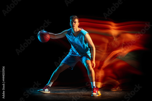 Young caucasian male basketball player playing basketball isolated over dark studio background in mixed light. Concept of healthy lifestyle, professional sport, hobby. © master1305
