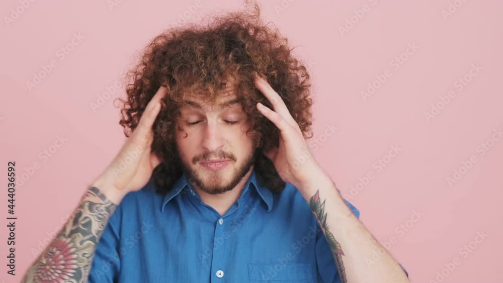 A nervous curly man wearing a blue shirt is grabbing his head isolated over pink background in the studio A nervous curly man wearing a blue shirt is grabbing his head isolated over pink background in the studio