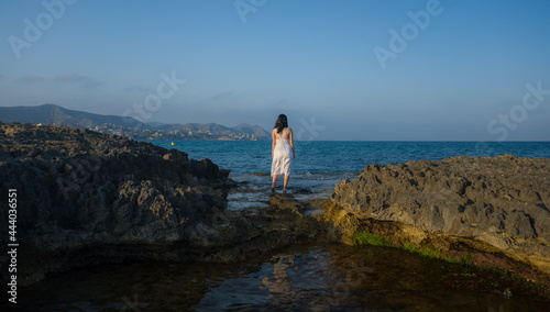 panoramic view of young attractive and relaxed woman from her back looking to the sea horizon wearing white summer dress enjoying beautiful holidays under a blue sky