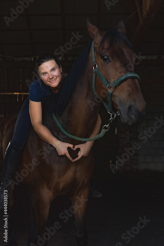 Horsewoman posing with her seal brown horse in the stable. Girl making a heart with her fingers. Expressing her love for the stallion. Bonding between human beings and horses concept. © CameraCraft