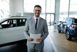 Confident young car dealer standing in showroom and holding a papers while posing.
