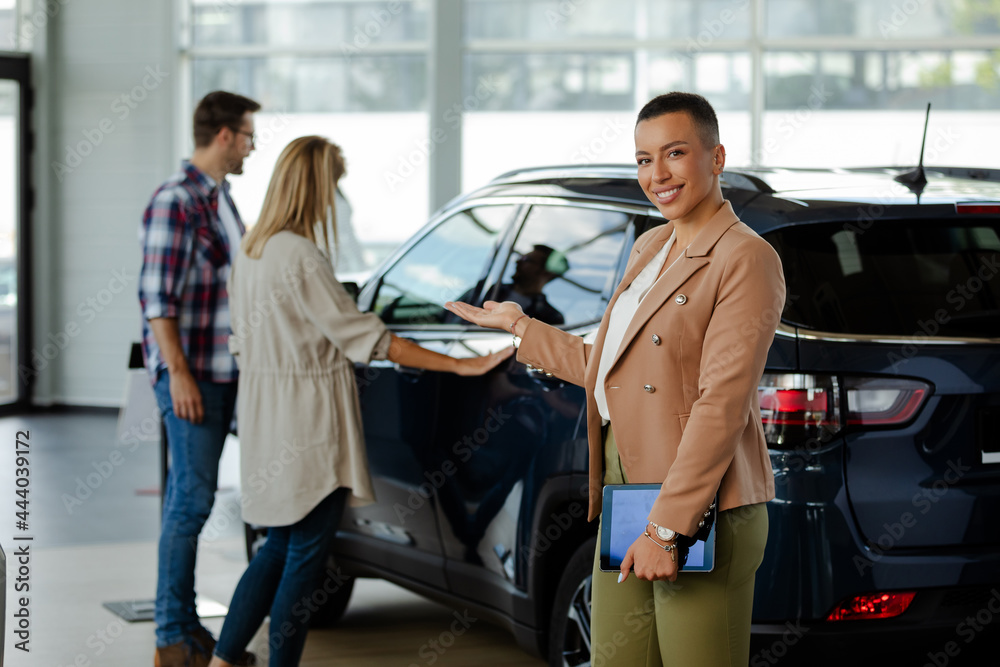 Saleswoman holding a tablet and showing on couple costumers while looking at camera at car showroom.
