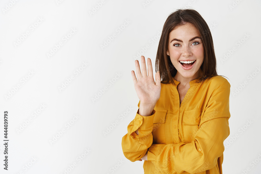 Friendly young woman waving hand and saying hi, greeting you with joyful smile, standing over white background Friendly young woman waving hand and saying hi, greeting you with joyful smile, standing over white background