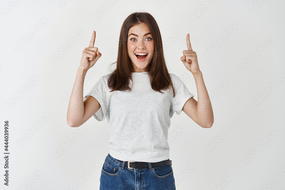 Surprised and happy woman showing advertisement, pointing hands up and looking at camera fascinated, standing over white background Surprised and happy woman showing advertisement, pointing hands up and looking at camera fascinated, standing over white background