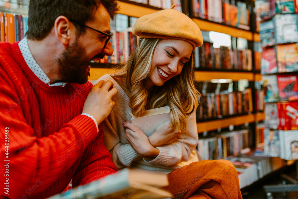Close up photo of laughing young couple sitting in bookstore. Young blonde woman is holding book on her chest while her boyfriend is sitting next to her.