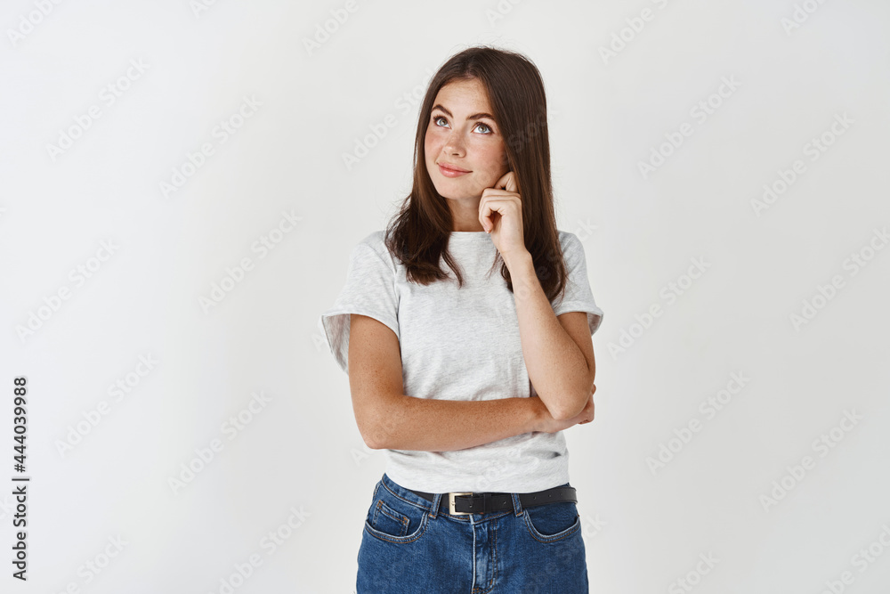 Thoughtful young woman making her decision, looking at upper left corner and smiling hesitant, standing over white background Thoughtful young woman making her decision, looking at upper left corner and smiling hesitant, standing over white background