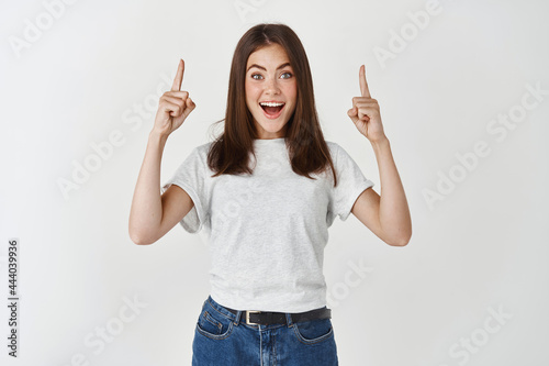 Surprised and happy woman showing advertisement, pointing hands up and looking at camera fascinated, standing over white background