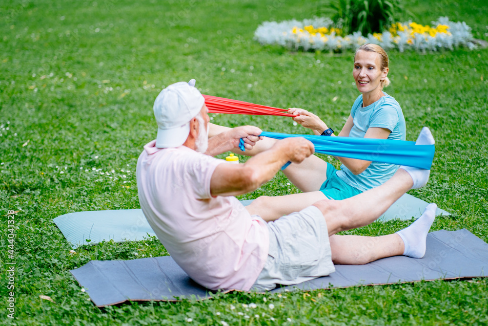 Active elderly man and young woman physiotherapist or instructor sitting on yoga mat in green grass meadow exercising with a rubber resistance band in green city park during his fitness workout. Active elderly man and young woman physiotherapist or instructor sitting on yoga mat in green grass meadow exercising with a rubber resistance band in green city park during his fitness workout.