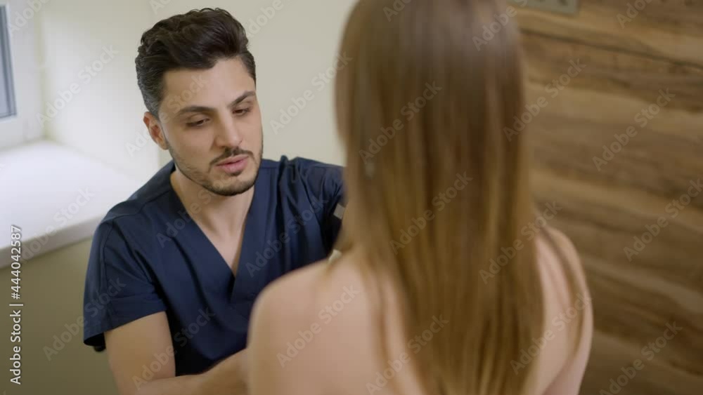 Serious concentrated Middle Eastern plastic surgeon checking breast of Caucasian patient in hospital talking in slow motion. Portrait of male expert consulting young woman on augmentation Serious concentrated Middle Eastern plastic surgeon checking breast of Caucasian patient in hospital talking in slow motion. Portrait of male expert consulting young woman on augmentation