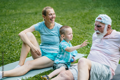 Happy family communicate together. Grandfather, mother and son sits on the green grass on mat together, relaxing after workout. Different generation. Family Vacation. Generational connection. © Iryna