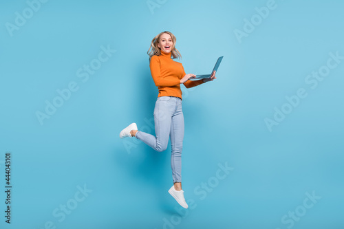 Photo of sweet excited young woman dressed orange pullover smiling jumping typing modern device isolated blue color background