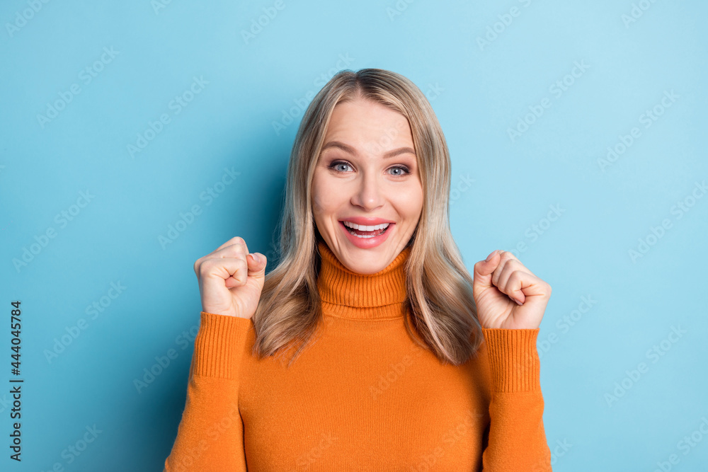Photo of cute lucky young lady wear orange sweater smiling rising fists isolated blue color background Photo of cute lucky young lady wear orange sweater smiling rising fists isolated blue color background