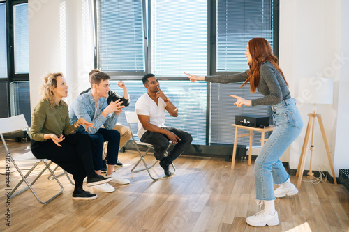 Fotografie Side view of cheerful redhead woman playing charades with friends showing pantomime by window in office