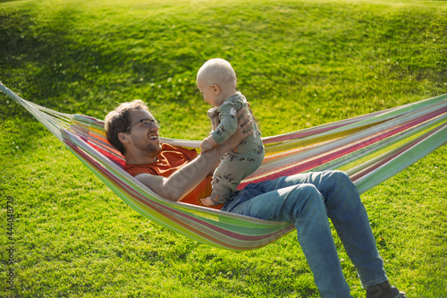 portrait of attractive nerd man with glasses in the park with green lawn have a nice sunset with a baby boy next to the hammock . Happy fatherhood
