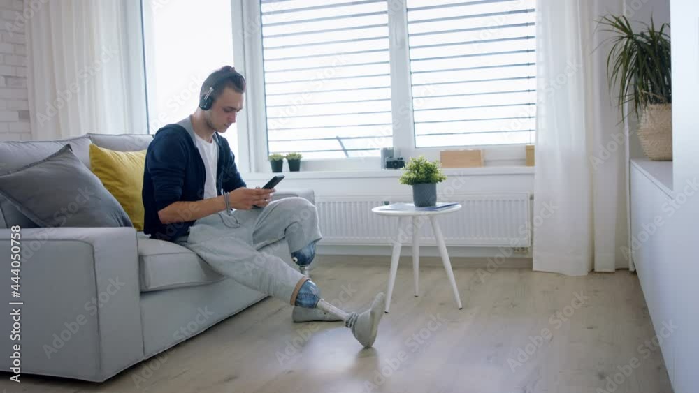 Disabled young man using smartphone indoors at home, leg prosthetic concept. Disabled young man using smartphone indoors at home, leg prosthetic concept.