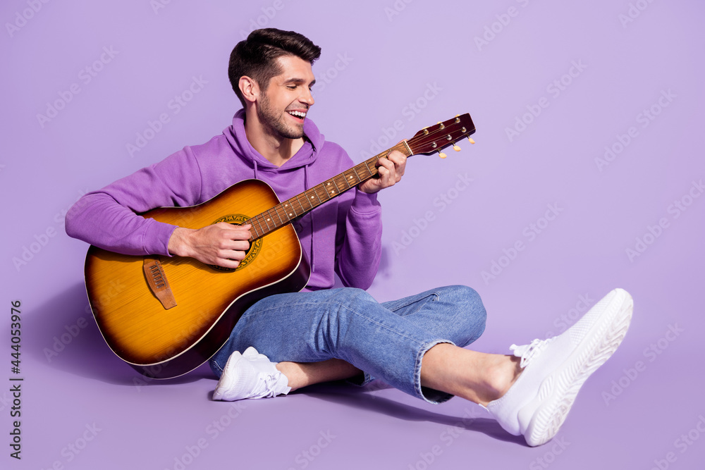 Full length body size photo man smiling sitting down playing guitar isolated pastel violet color background Full length body size photo man smiling sitting down playing guitar isolated pastel violet color background