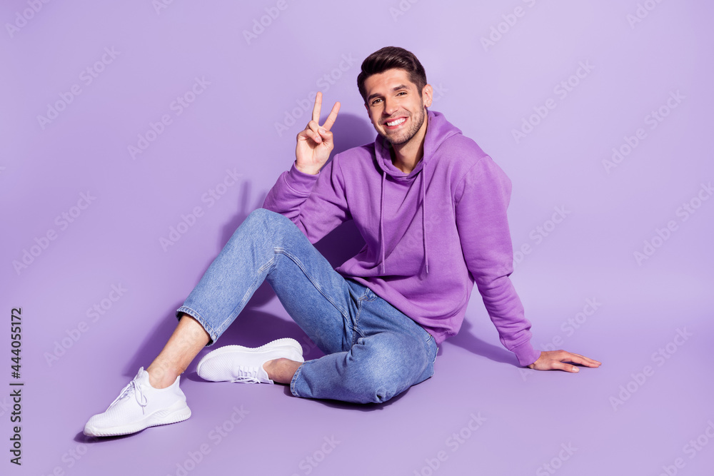 Portrait of attractive cheerful guy sitting showing v-sign good mood isolated over purple violet color background Portrait of attractive cheerful guy sitting showing v-sign good mood isolated over purple violet color background