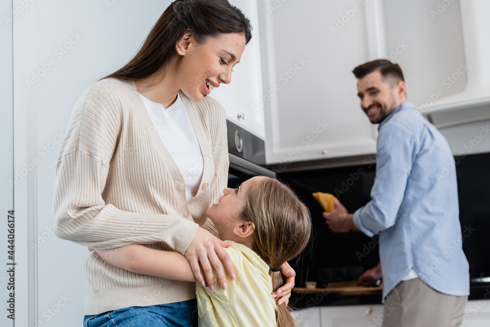 Fototapeta premium excited woman embracing daughter near blurred husband holding bread in kitchen