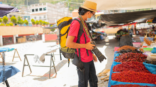 A tourist with a medical mask, backpack, and professional camera, walks in the middle of the sales of a local market.
