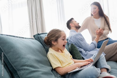 excited girl using laptop near happy parents holding hands and talking on blurred background
