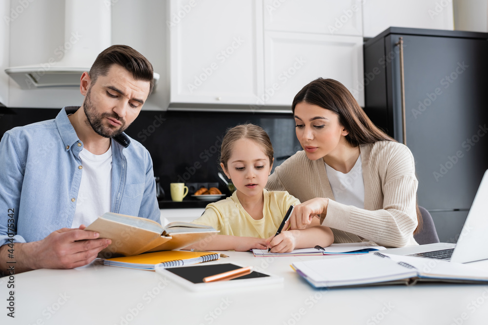 man reading book and woman pointing with finger while helping daughter doing homework man reading book and woman pointing with finger while helping daughter doing homework
