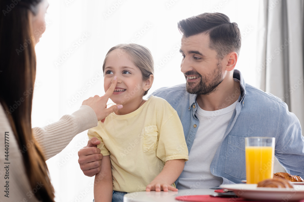 mother touching nose of cheerful daughter near happy husband during breakfast mother touching nose of cheerful daughter near happy husband during breakfast