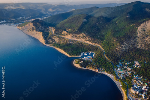 Aerial view of coastline with blue sea, mountains with trees, cliffs with sunset light.