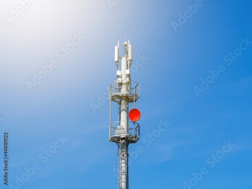 A 5G telecommunications tower on the background of a clear blue sky without clouds. A gray cellular antenna with a red satellite dish. A wireless communication base station.