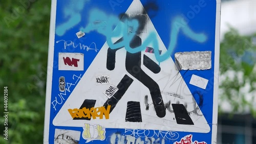 Vandalized pedestrian zebra crossing sign painted by vandals. Metallic road sign with graffiti and funny stickers. Damaged crosswalk banner. Problem of street vandalism in urban environment