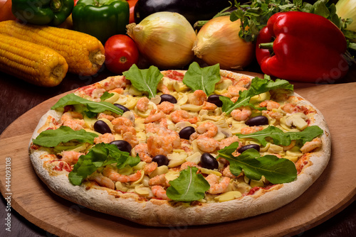 Shrimp and mushroom pizza with arugula on a wooden board and vegetables in the background.