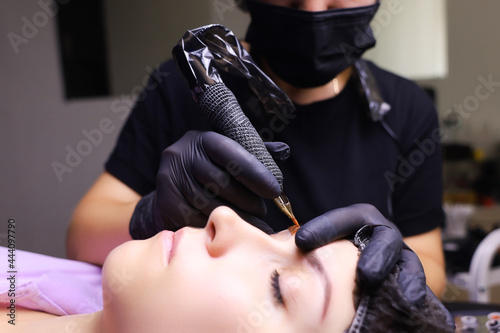 tattoo artist in black gloves with a tattoo machine for applying pigment to the client s eyebrows