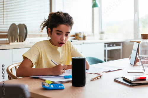 Hispanic girl doing homework and talking to smart speaker photo