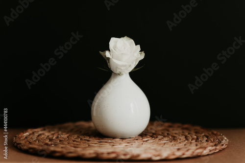 A white rose head in a white ceramic vase on brown woven place mat and black background. photo