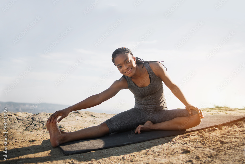 Smiling fitness afro-american woman in sport clothes stretching body on yoga mat outdoors. Slender female having morning workout on fresh air. Active and healthy lifestyles.