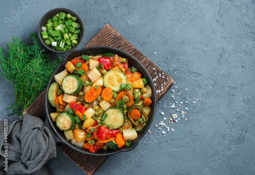 Frying pan with fried vegetables on a wooden board on a gray-blue background.